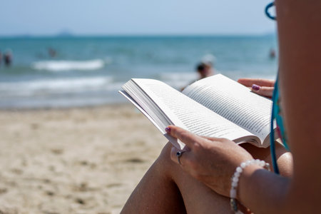 Woman reading a book on the beach, blurred background. Summer leisure on the beach.