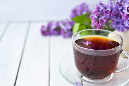 Delicate setting of the morning tea table with lilac flowers, a transparent cup of tea and a saucer and a white vase on a white wooden board. Copy space. Spring breakfast concept.の写真素材