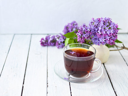 Delicate setting of the morning tea table with lilac flowers, a transparent cup of tea and a saucer and a white vase on a white wooden board. Copy space. Spring breakfast concept.の写真素材