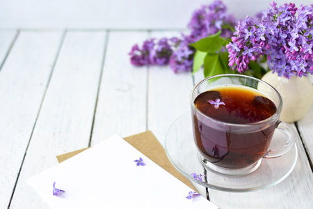 Delicate morning tea table setting with lilac flowers, a transparent cup of tea and a saucer and a white vase, white and craft note paper on a white wooden board. Mockup. Copy space. Spring breakfast concept.の写真素材