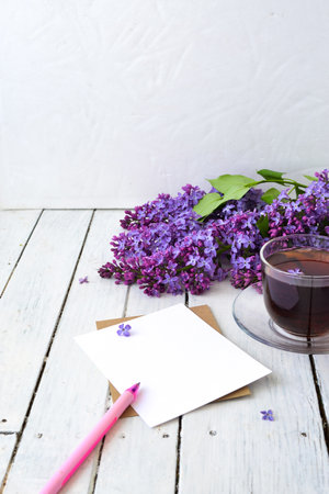 Delicate morning tea table setting with lilac flowers, a transparent cup of tea and a saucer and a white vase, white and craft paper for notes, and a pink pen on a white wooden table. Mockup. Copy space. Spring breakfast concept.の写真素材