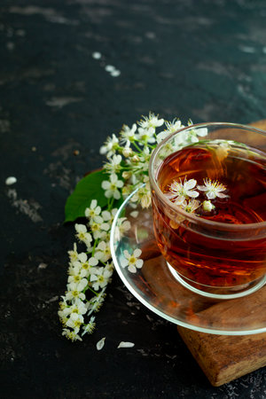 A transparent cup of teapot tea with a saucer and a white bird cherry flower on a black background. Copy space. Spring breakfast.の写真素材