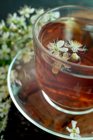 A transparent cup of teapot tea with a saucer and a white bird cherry flower on a black background. Copy space. Spring breakfast.の写真素材