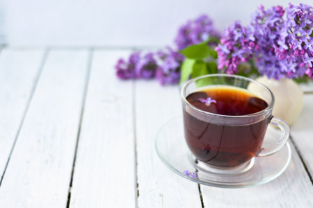 Delicate setting of the morning tea table with lilac flowers, a transparent cup of tea and a saucer and a white vase on a white wooden board. Copy space. Spring breakfast concept.の写真素材