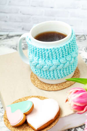 A white cup of tea in a knitted blue case stands on a book and pink flowers, tulips and gingerbread on a texture table. Spring concept.の写真素材