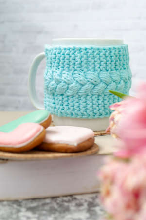 A white cup of tea in a knitted blue case stands on a book and pink flowers, tulips and gingerbread on a texture table. Spring concept.の写真素材