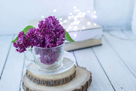 Transparent cup with flowers. Beautiful lilac fills the glass. The spring bouquet is worth no wooden cuts. Macro image of spring lilac purple flowers. Book in the background and lights. Blurredの写真素材