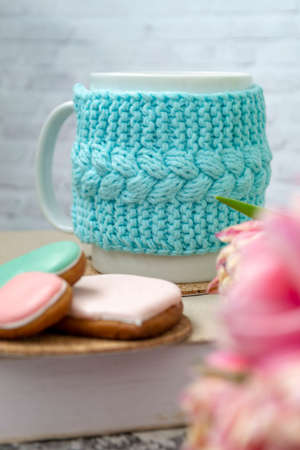 A white cup of tea in a knitted blue case stands on a book and pink flowers, tulips and gingerbread on a texture table. Spring concept.の写真素材