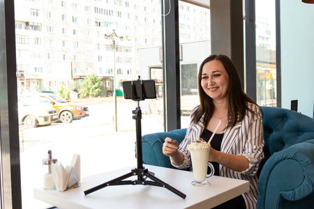 A young laughing brunette sits at a table in a cafe. She speaks by video communication on the phone, and on the table is a glass with a cocktail. She communicates via video link with a friend. Communication at a distanceの写真素材