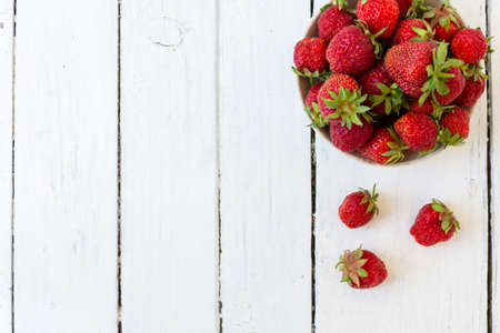 Ripe red strawberries lie in a white cup on a wooden white table. Copy space. View from above