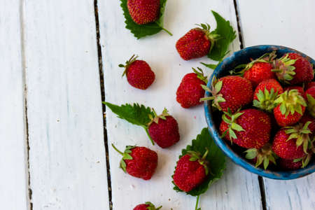 Ripe red strawberries lie in a blue cup on a wooden white table. Sweet berries are scattered nearby. Copy space. top view of the photoの写真素材