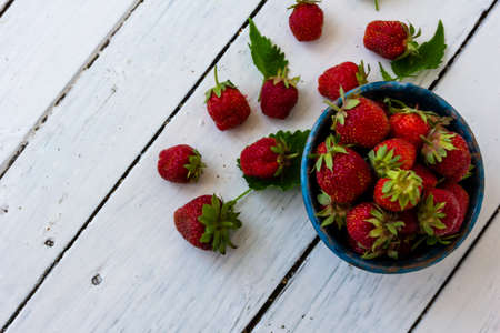 Ripe red strawberries lie in a blue cup on a wooden white table. Sweet berries are scattered nearby. Copy space. top view of the photo.の写真素材