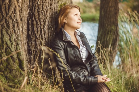 beautiful girl Asian girl sits near a tree on the grass in the jacketの写真素材