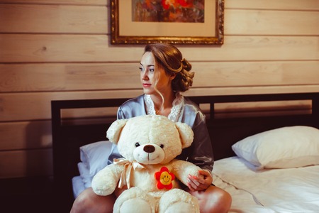 portrait of cute young girl who is sitting on the bed in his room and holds a soft toy in the hands and smilingの写真素材