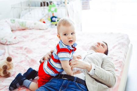 Happy father plays with his son sitting on the bed at home. Young man playing with little boy.