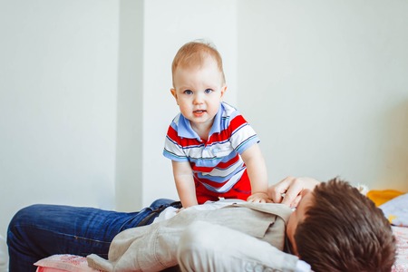 Happy father plays with his son sitting on the bed at home. Young man playing with little boy.の写真素材