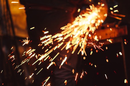 Craftsman sawing metal sparkles all around workshop. Working at noon.