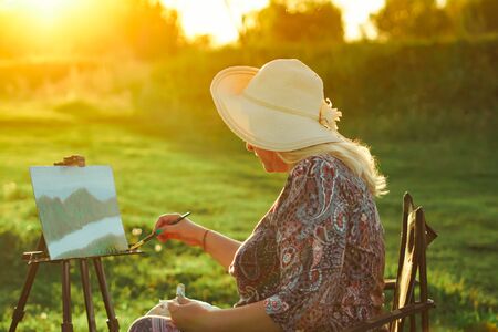 an elderly woman paints a picture in nature oil painting by the riverの写真素材