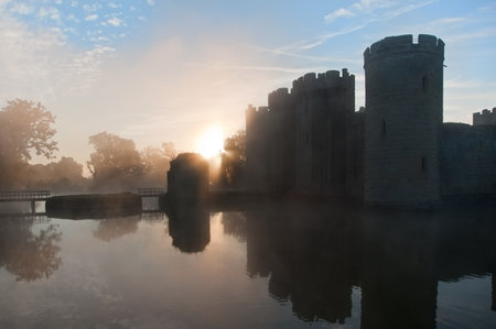 Beautiful medieval castle and moat at sunrise with mist over moat and sunlight behind castleのeditorial素材