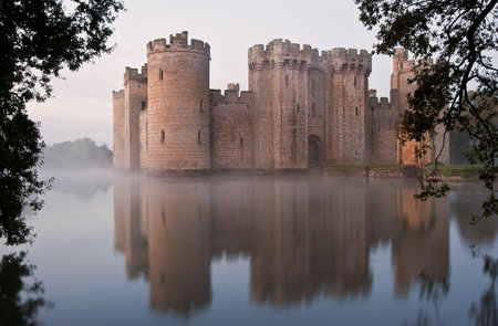 Beautiful medieval castle and moat at sunrise with mist over moat and sunlight behind castleのeditorial素材