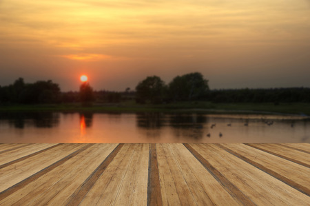Beautiful simple image of sunset through tress reflected in lake in foreground with wooden planks floorの写真素材