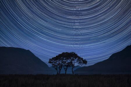 Digital composite image of star trails around Polaris with beautiful foggy misty countryside landscape surrounding crummock water in lake district in englandの写真素材
