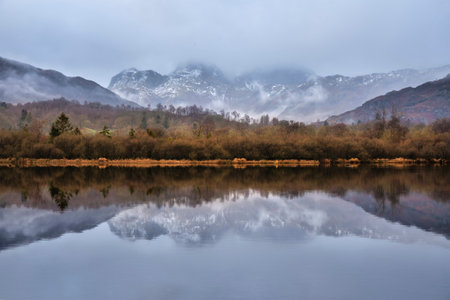 Epic dramatic landscape image looking across River Brathay in Lake District towards Langdale Piks mountain range on mistry Winter morningの写真素材