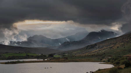 Epic Autumn landscape image of Snowdon Massif viewed from shores of Llynnau Mymbyr at sunset with dramatic dark sky and cloudsの写真素材