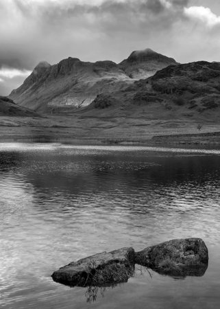 Black and white Epic landscape of sunrise light over Blea Tarn in Lake District with stunning light on distant mountainsの写真素材