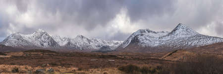 Stunning Winter panorama landscape image of mountain range viewed from Loch Ba in Scottish Highlands with dramatic clouds overheadの写真素材