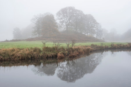 Epic Autumn landscape image of River Brathay in Lake District lookng towards Langdale Pikes with fog across river and vibrant woodlandsの写真素材