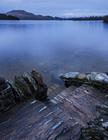 Stunning landscape image of Loch Lomond and snowcapped mountain range in distance viewed from small village of Lussの写真素材