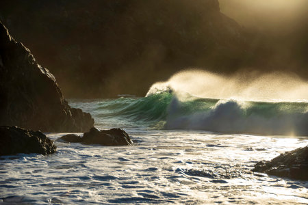Epic landscape image of jade turquoise waves crashing onto shore and rocks in Kynance Cove Cornwall with glowing sunrise background and water spray droplets in windの写真素材
