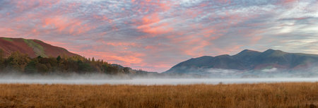 Epic Autumn sunrise landscape image looking from Manesty Park in Lake Distict towards sunlit Skiddaw Range with mit rolling across Derwentwater surfaceの写真素材