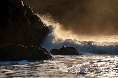 Epic landscape image of jade turquoise waves crashing onto shore and rocks in Kynance Cove Cornwall with glowing sunrise background and water spray droplets in windの写真素材