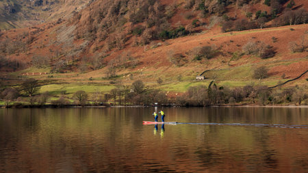 Beautiful Winter landscape views of mountain ranges around Ullswater in Lake District viewed from boat on lake with unidentified people paddle boarding on lakeの写真素材