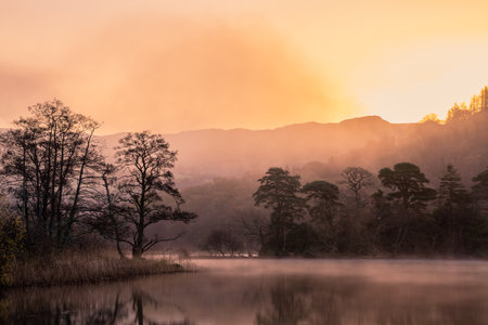 Dramatic atmospheric sunrise landscape at Rydal Water in Lake District during Autumn with moody glow...