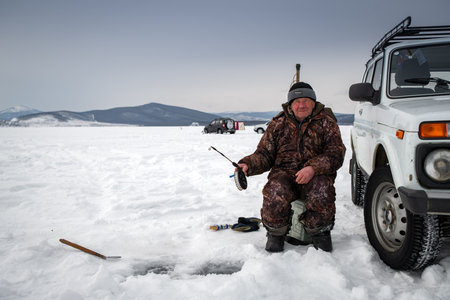 Baikal lake, Siberia, Russia - March 05, 2020 : Winter ice fishing. A fisherman is fishing on spinings near tents and cars on Lake Baikal on a March day. Baikal lake, Siberia, Russiaのeditorial素材