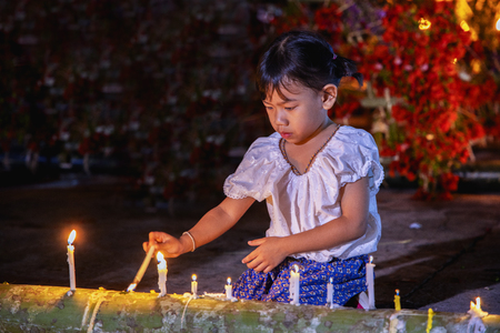 Pretty small girl is looking on the light of candle. Little girl holds a burning church candle and reads a prayer.The little girl is burning candles in the flower parade in the temple of Thailand.の写真素材