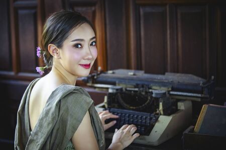 A beautiful Thai girl wearing a traditional Thai dress is using an old typewriter and looking at the camera. Retro typewriter placed on wooden planks.の写真素材