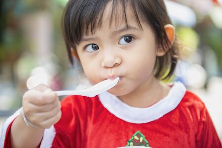 The cute little girl is eating food with a plastic spoon.の写真素材