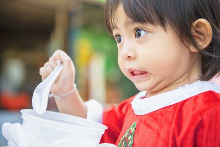 The cute little girl is eating food with a plastic spoon. Cute little Asian girl wear Christmas costumes to celebrate Christmas.の写真素材