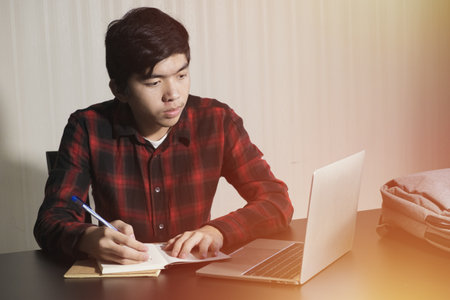 Learn online, Student watching lesson online and studying from home. Young man taking notes while looking at computer screen following professor on video call. Studying from home on pc.の写真素材