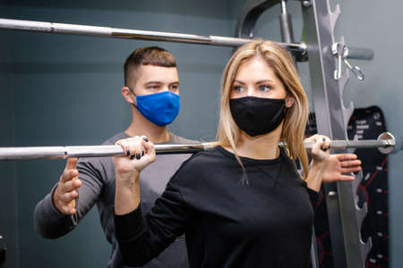 Young woman with protective mask working out with personal trainer at the gym during pandemic. She...