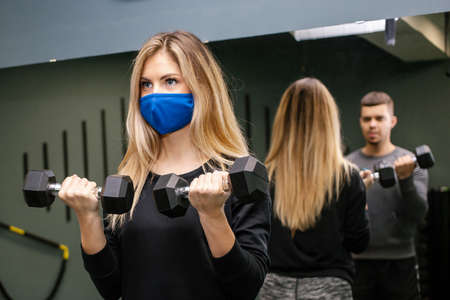 Young pretty woman with protective mask working out with personal trainer at the gym during ...