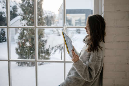 Young cute woman in cozy sweater sitting in bedroom and reading a book near window.の写真素材