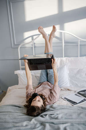 Creative woman is lying on bed with laptop, smiling thoughtful, writes a message, calls via video chat, reads the news. Work from home, freelance and lifestyle concept.の写真素材