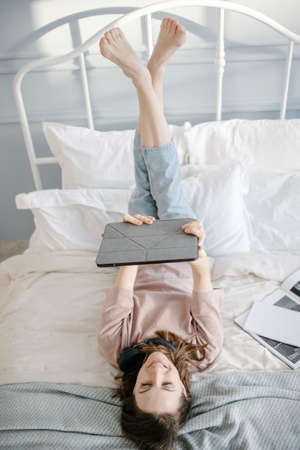 Creative woman is lying on bed with tablet, smiling thoughtful, writes a message, calls via video chat, reads the news.の写真素材