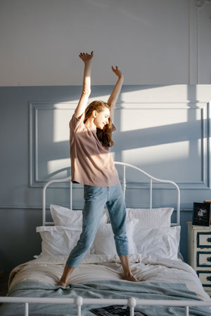 Happy young beautiful woman pulls her hands up, standing on the bed in her bedroom at home. People and Morning concept.の写真素材