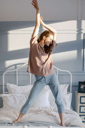 Happy young beautiful woman pulls her hands up, standing on the bed in her bedroom at home. People and Morning concept.の写真素材